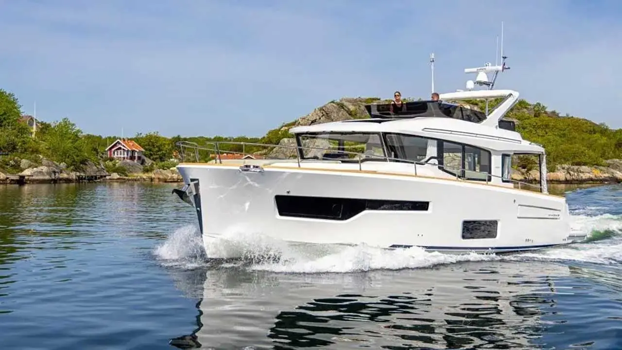 White motor yacht cruising through calm coastal waters, with rocky islands and a red cottage in the background.