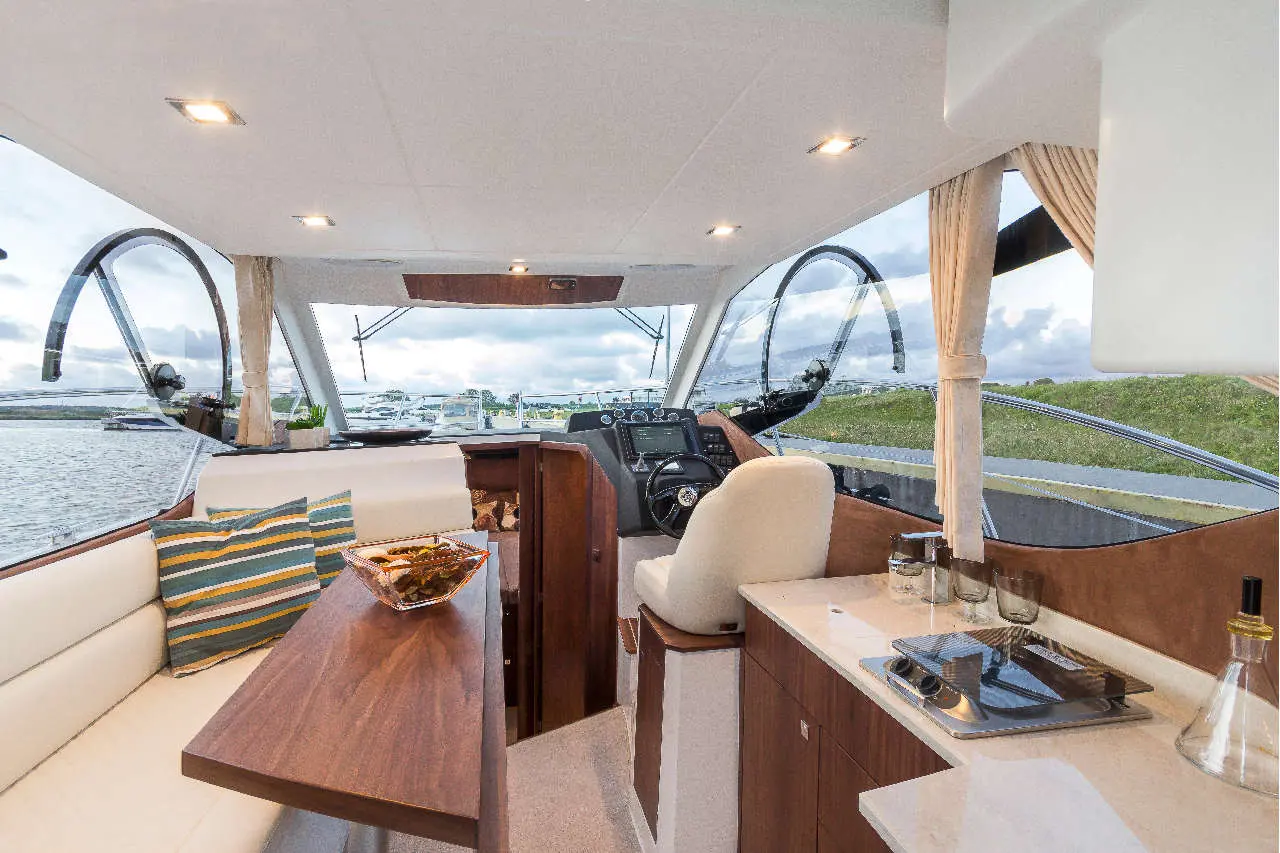 Modern boat interior featuring a dining area, steering controls, and a view of the harbor with yachts and a cloudy sky.
