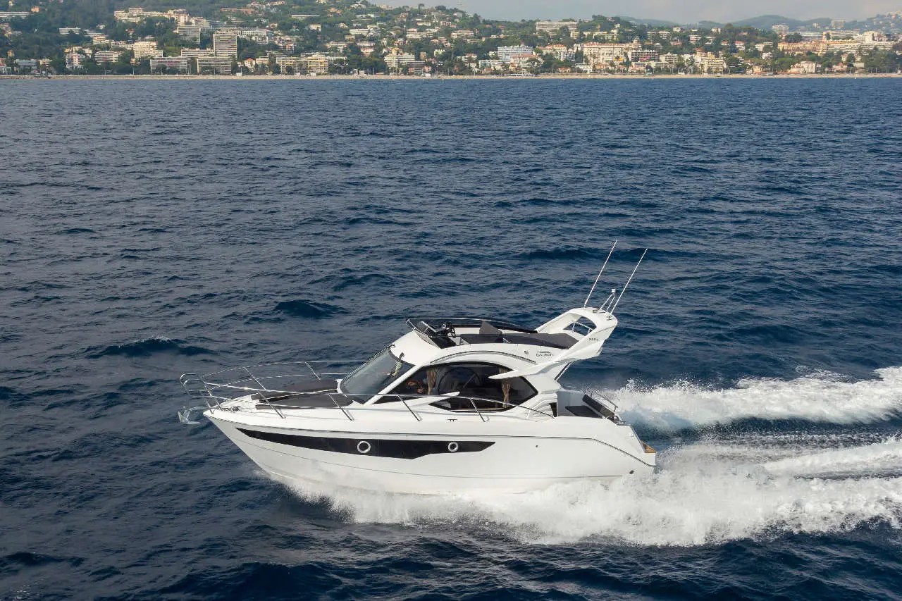A sleek white motor yacht speeds through blue waters with a coastal city in the background, showcasing a scenic maritime view.