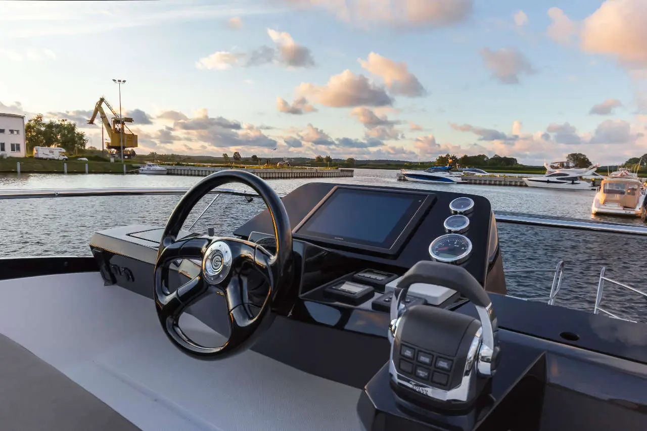 Boat cockpit view with steering wheel, control panel, and waterway; boats in the background under a cloudy sky.