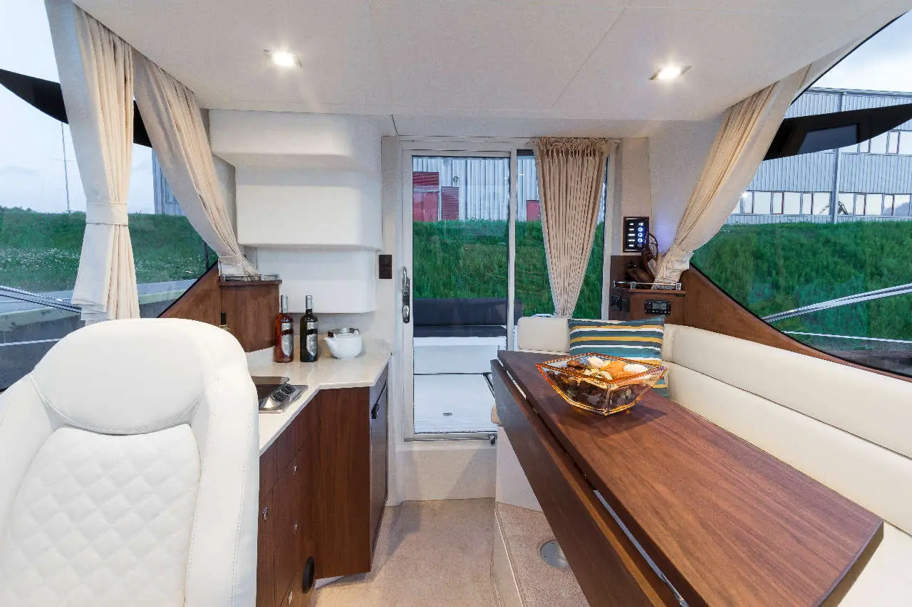 Modern yacht interior featuring a dining area, kitchen space, and large windows with natural light. Bottles and decor visible.