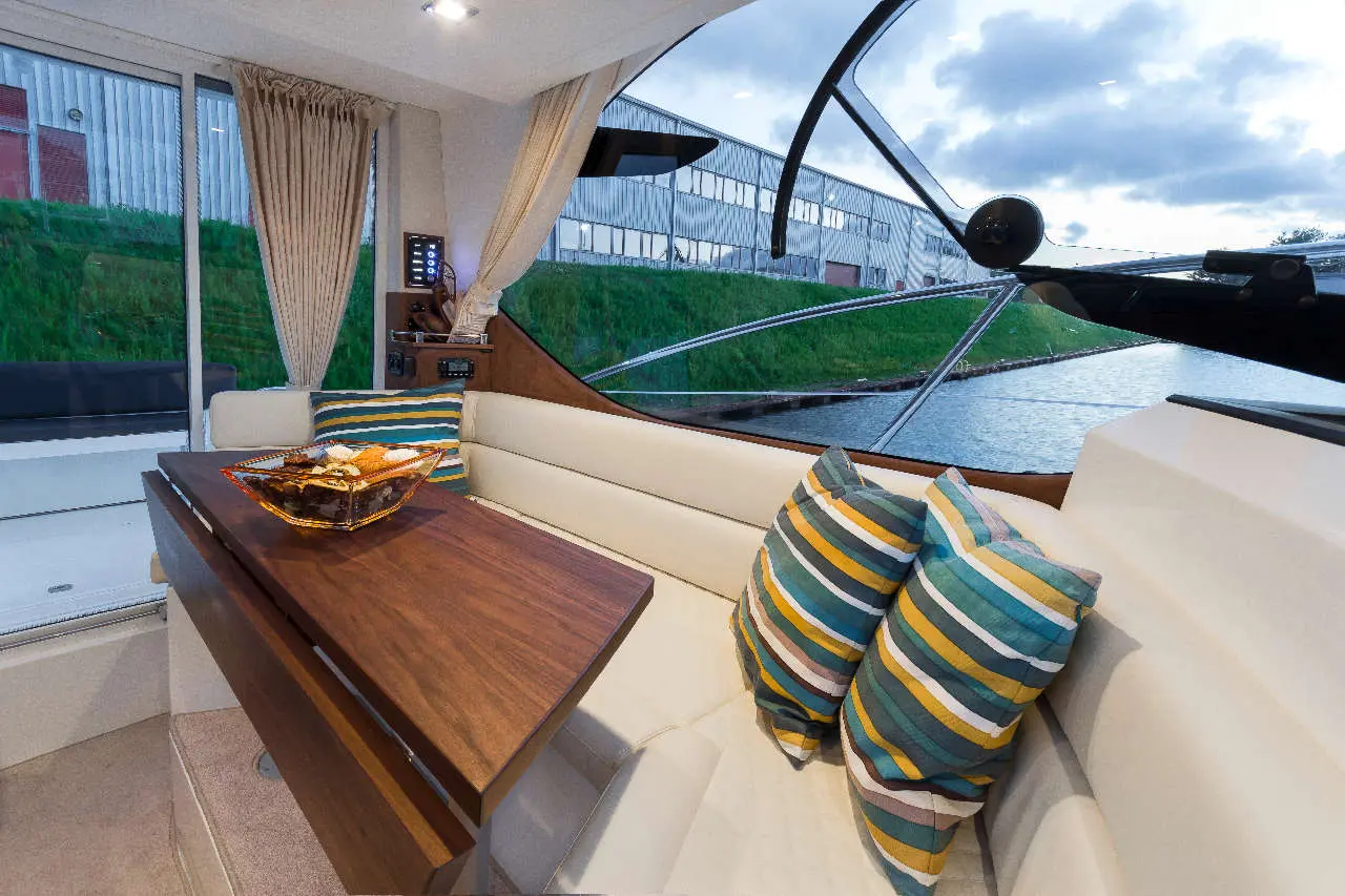 Interior of a boat featuring a wooden table, cream seating, striped cushions, and a view of the water and greenery outside.