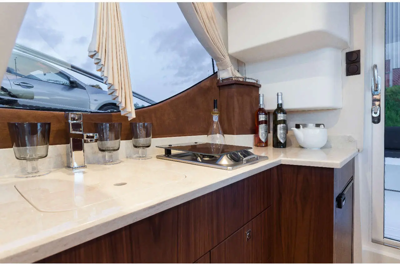 Stylish boat kitchenette featuring a sink, cooking hob, glasses, wine bottles, and large window with curtains.
