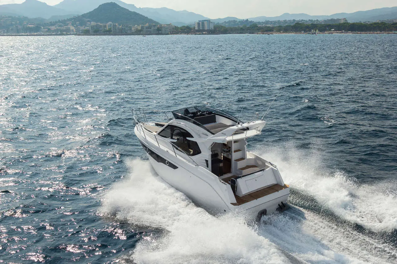 A white motorboat speeds across shimmering waters, with a distant coastal landscape and mountains visible in the background.