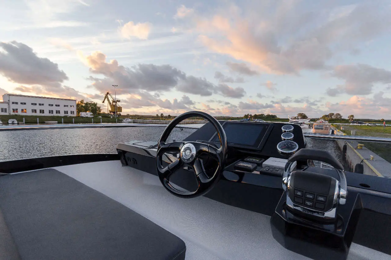 Boat cockpit with a steering wheel, control panel, and scenic waterfront view at sunset with clouds.