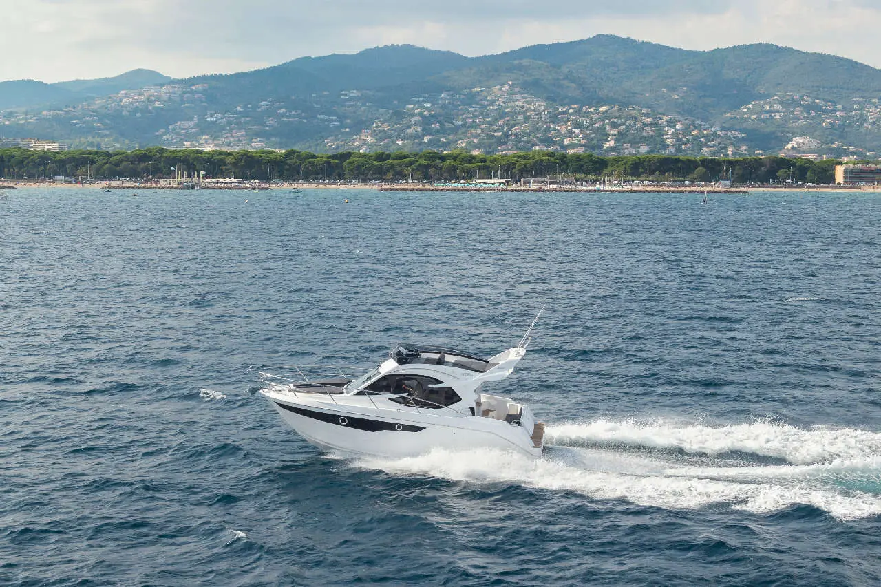 A white motorboat speeds across the calm sea near a sandy beach and green hills under a partly cloudy sky.