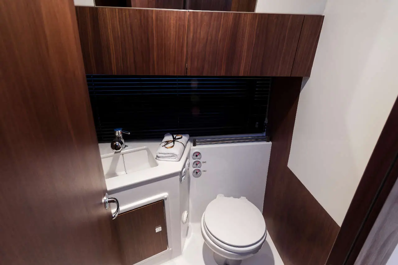 Compact bathroom with wood paneling, a toilet, sink, and towels on the counter, featuring blinds for privacy.