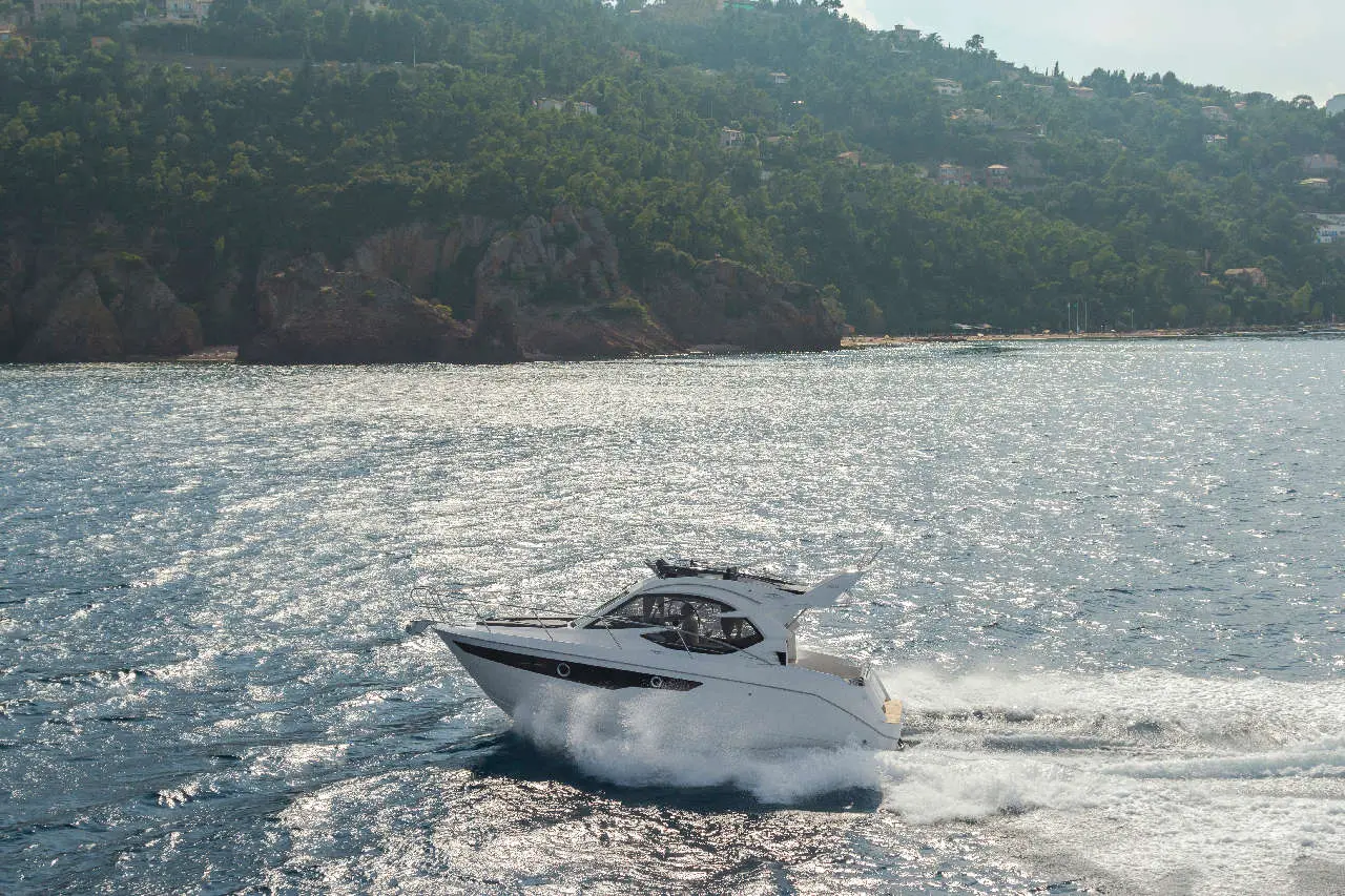 A white yacht speeds across shimmering water near a lush green coastline with rocky cliffs and scattered houses.