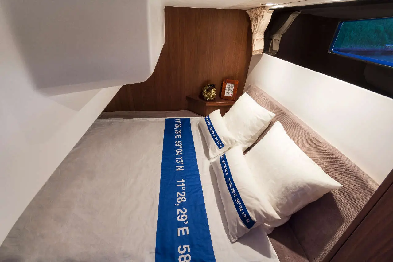 Compact bedroom with a small double bed, two pillows, and blue bed linen featuring GPS coordinates. Warm wooden accents visible.