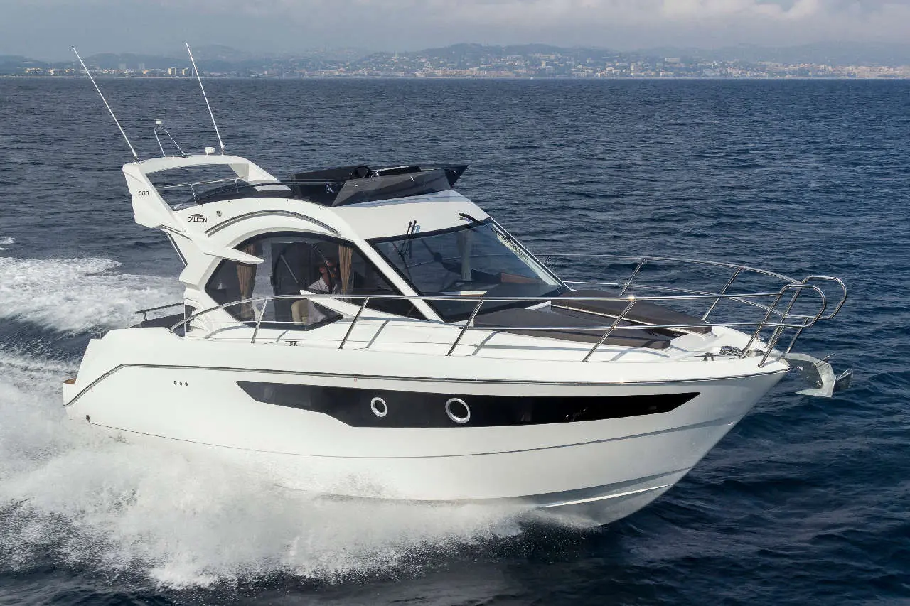 A modern motor yacht cruises swiftly through blue waters with a driver visible at the helm, coastal buildings in the distance.