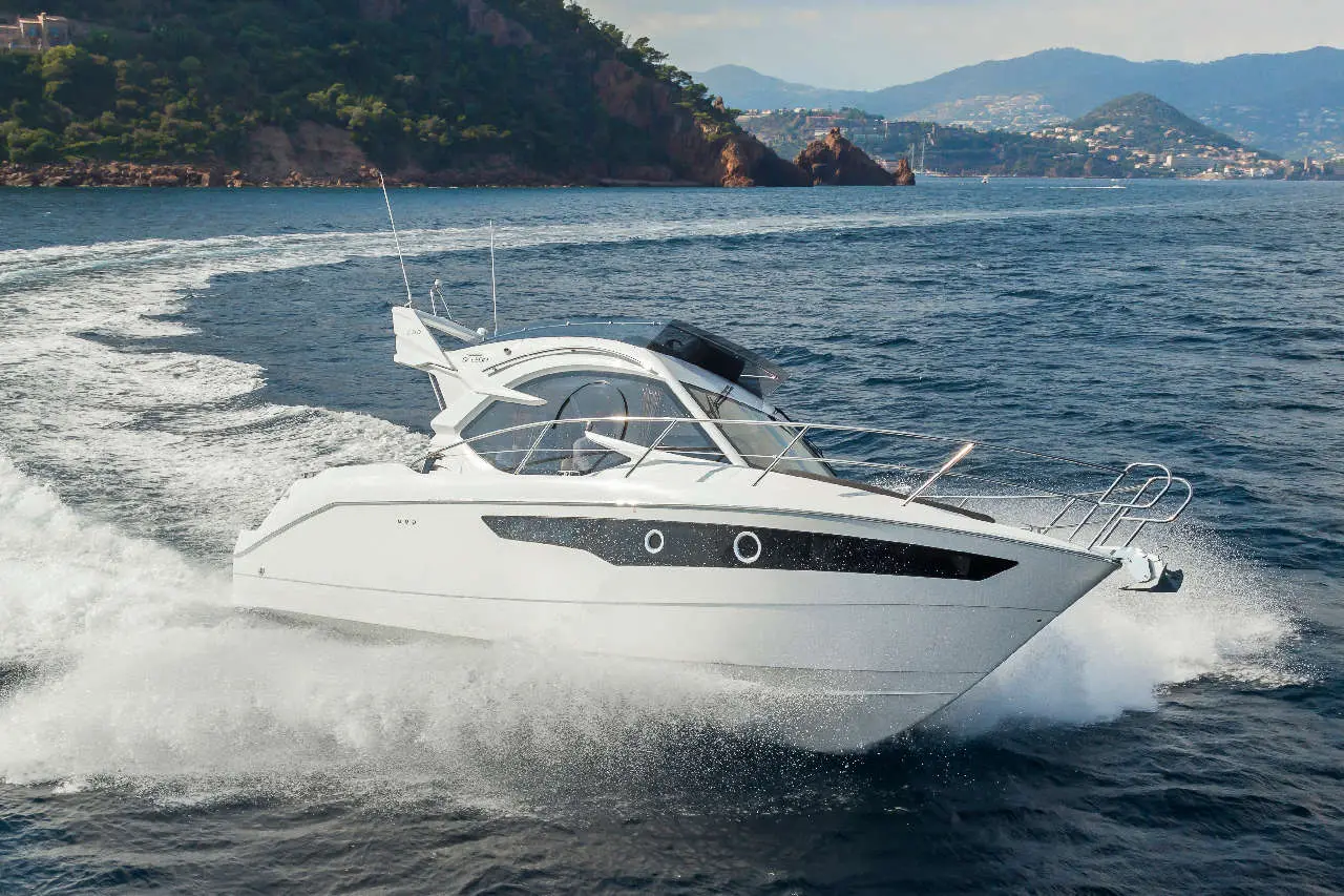 A white motorboat cruising on the sea, creating waves with a backdrop of green hills and distant coastline in sunlight.