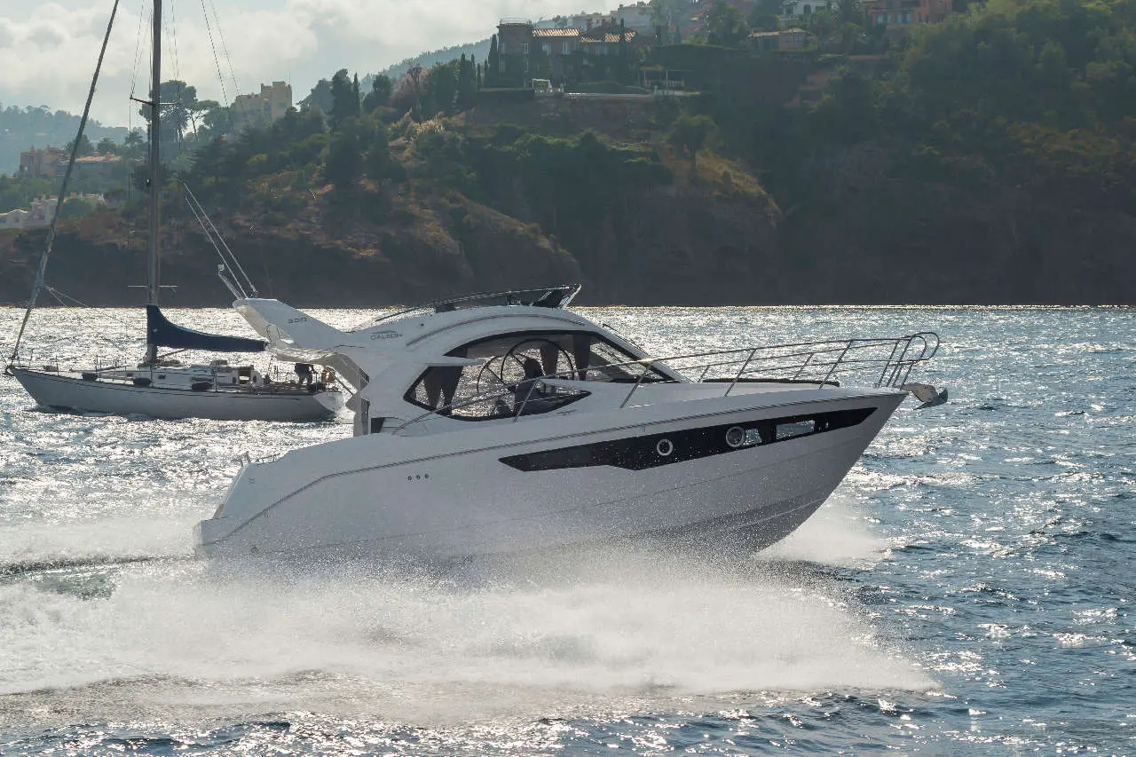 White motorboat splashes through water beside a sailboat, with coastal hills and homes in the background.