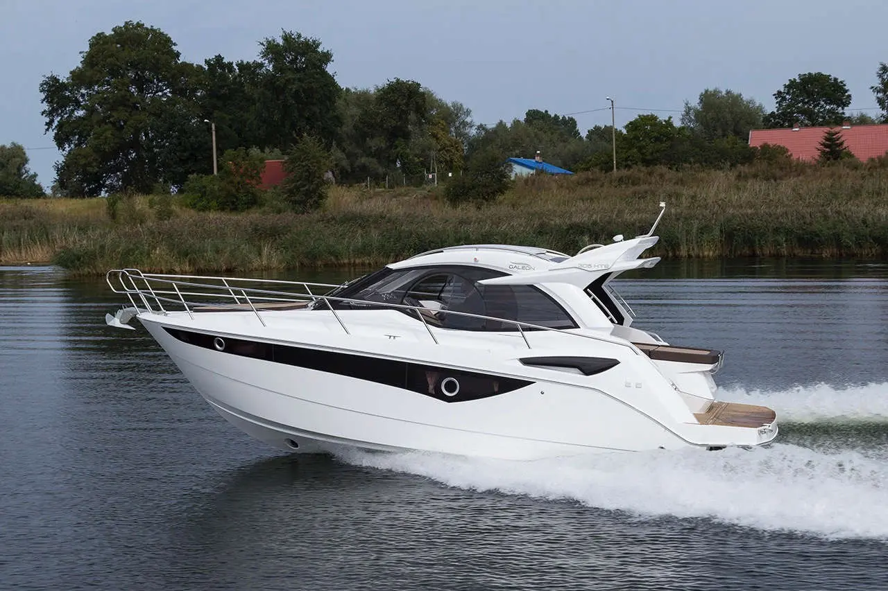 A sleek white motorboat glides across calm water, leaving a wake, surrounded by lush greenery and distant buildings.