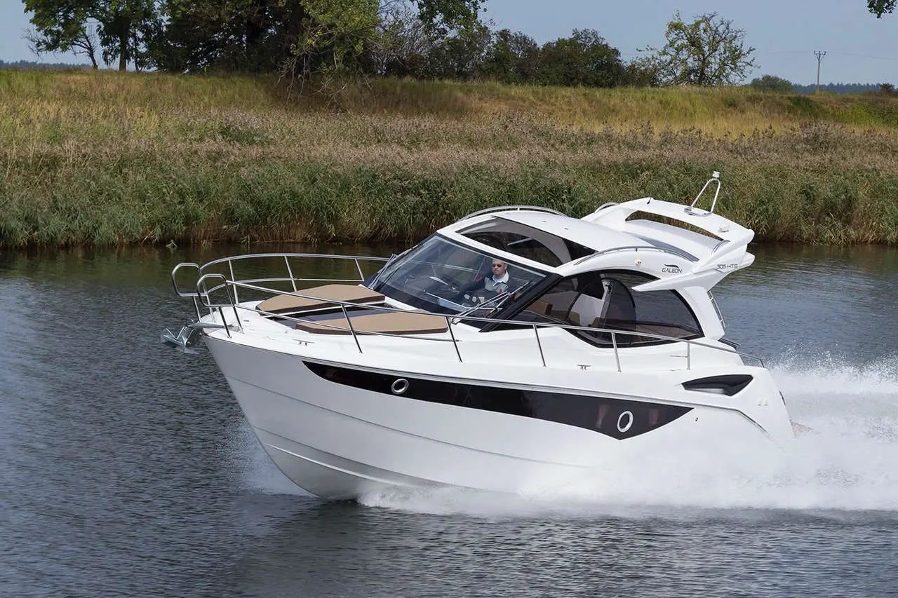 A sleek white motorboat speeds across calm water, with a driver behind the wheel surrounded by lush greenery.