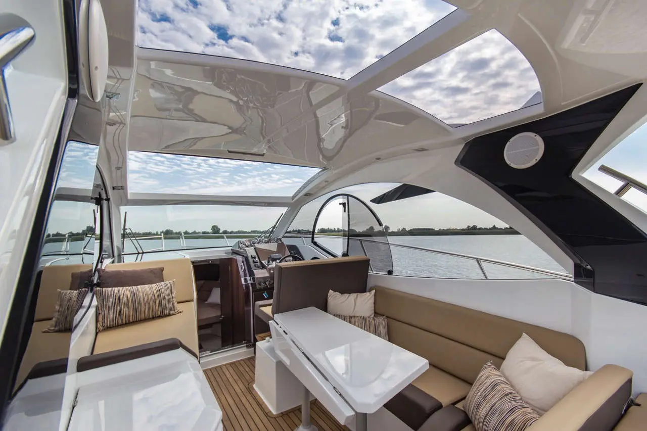 Luxury boat interior featuring modern seating, table, and large windows with scenic water view and clouded sky.