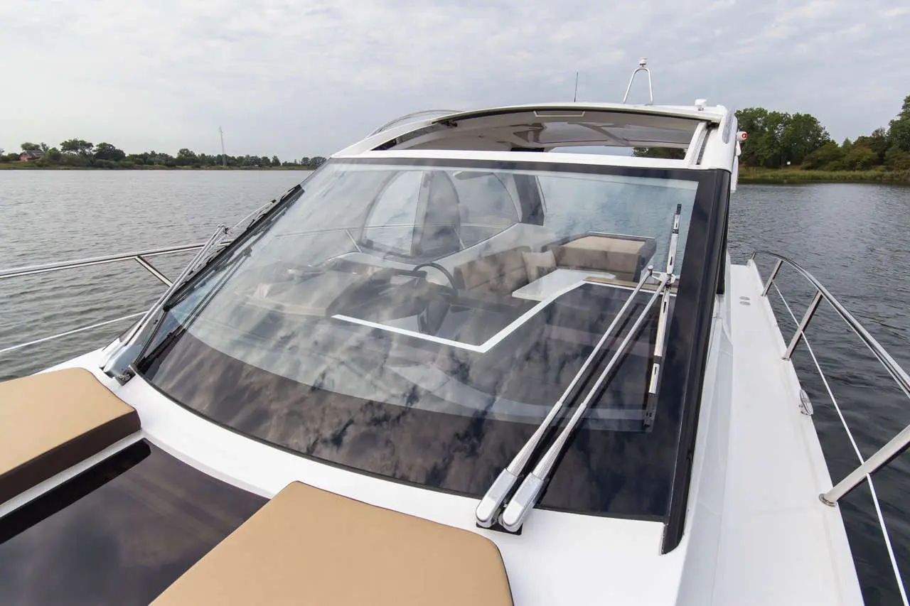 A luxury yacht's cockpit view with a large windshield, plush seating, and a scenic river backdrop under a cloudy sky.