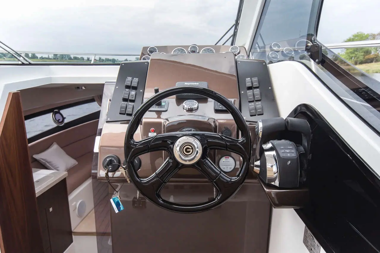 Boat cockpit with a black steering wheel, control panels, gauges, and soft seating in the background. Power and navigation controls visible.
