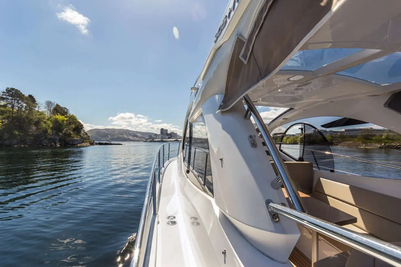 A yacht cruising through calm waters, with views of greenery and buildings under a clear blue sky.