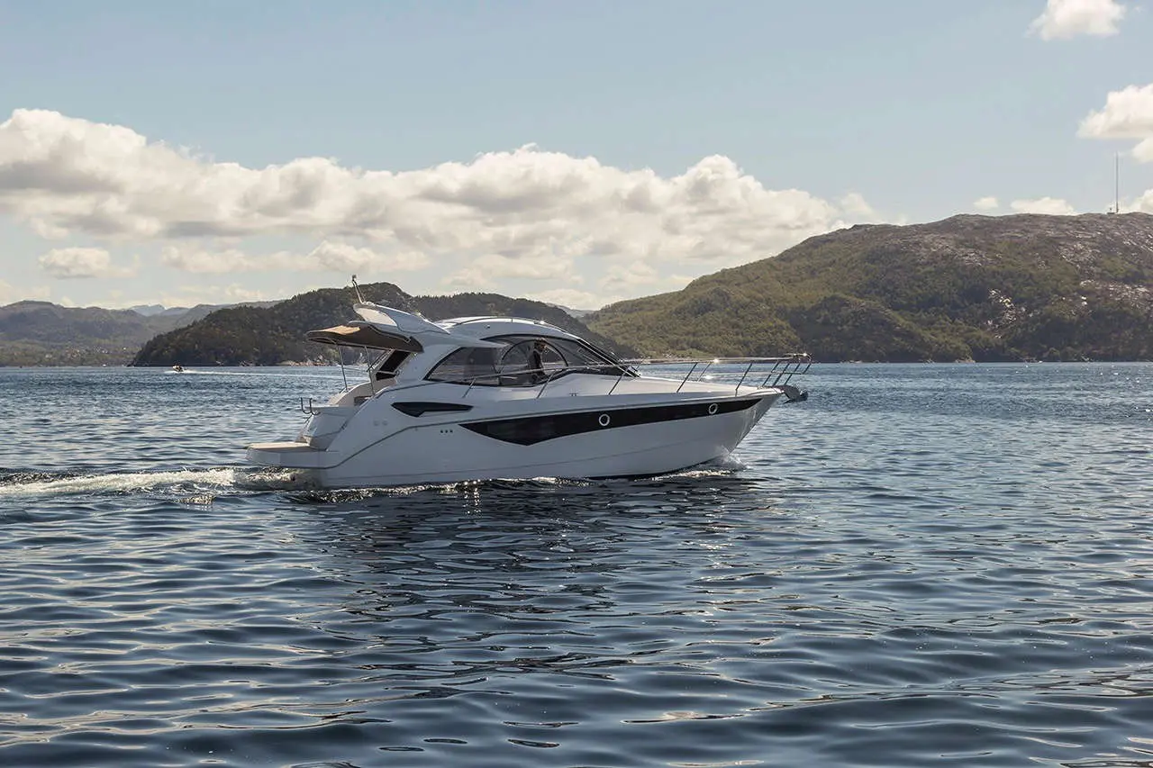 A sleek white motorboat glides through calm waters, surrounded by hills under a bright blue sky with fluffy clouds.