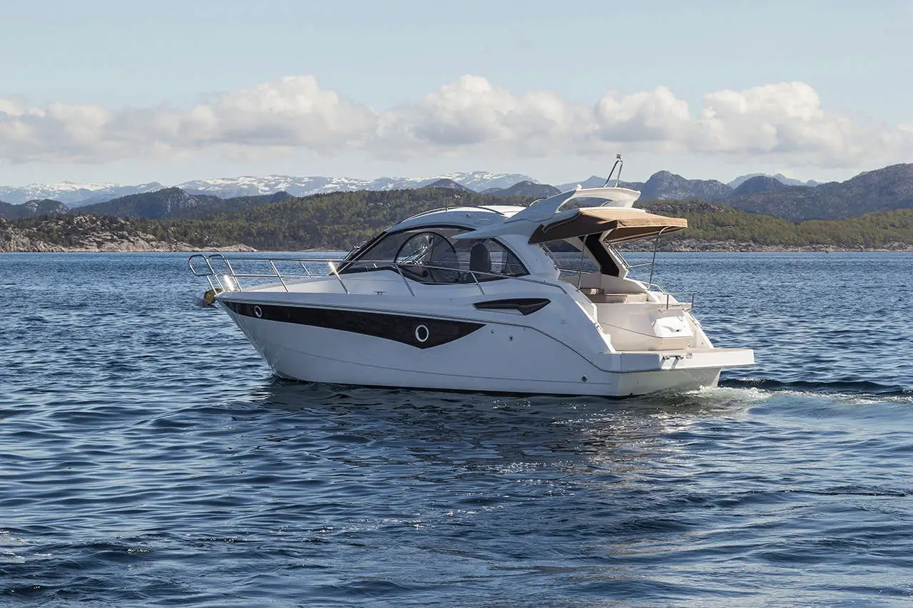 A sleek white motorboat cruising through calm water, with snow-capped mountains and a blue sky in the background.