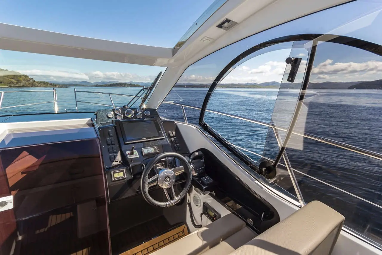 Modern yacht interior with a steering wheel, control panel, and panoramic views of water and hills under a clear blue sky.