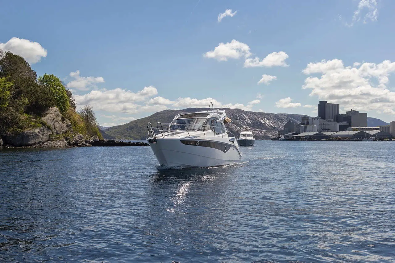 A white motorboat navigates calm waters, surrounded by green hills and industrial buildings under a blue sky with clouds.