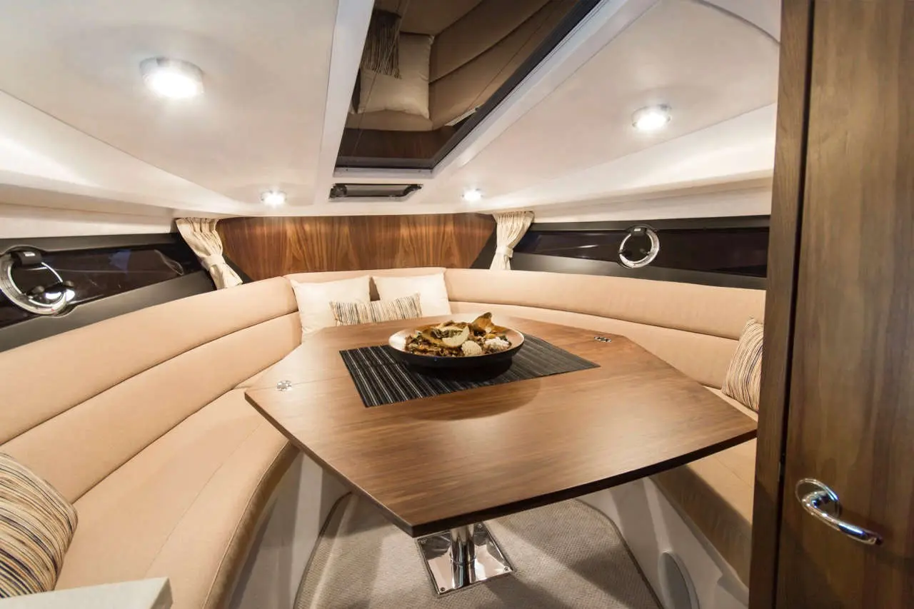 Interior of a boat featuring a circular dining area with a wooden table, cushions, and a platter of food on a black mat.