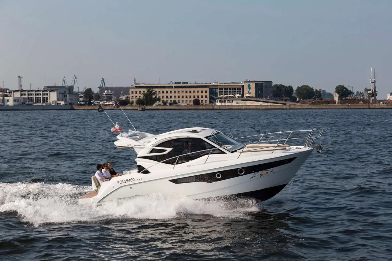 A white motorboat with two passengers speeds across the water, city buildings visible in the background.