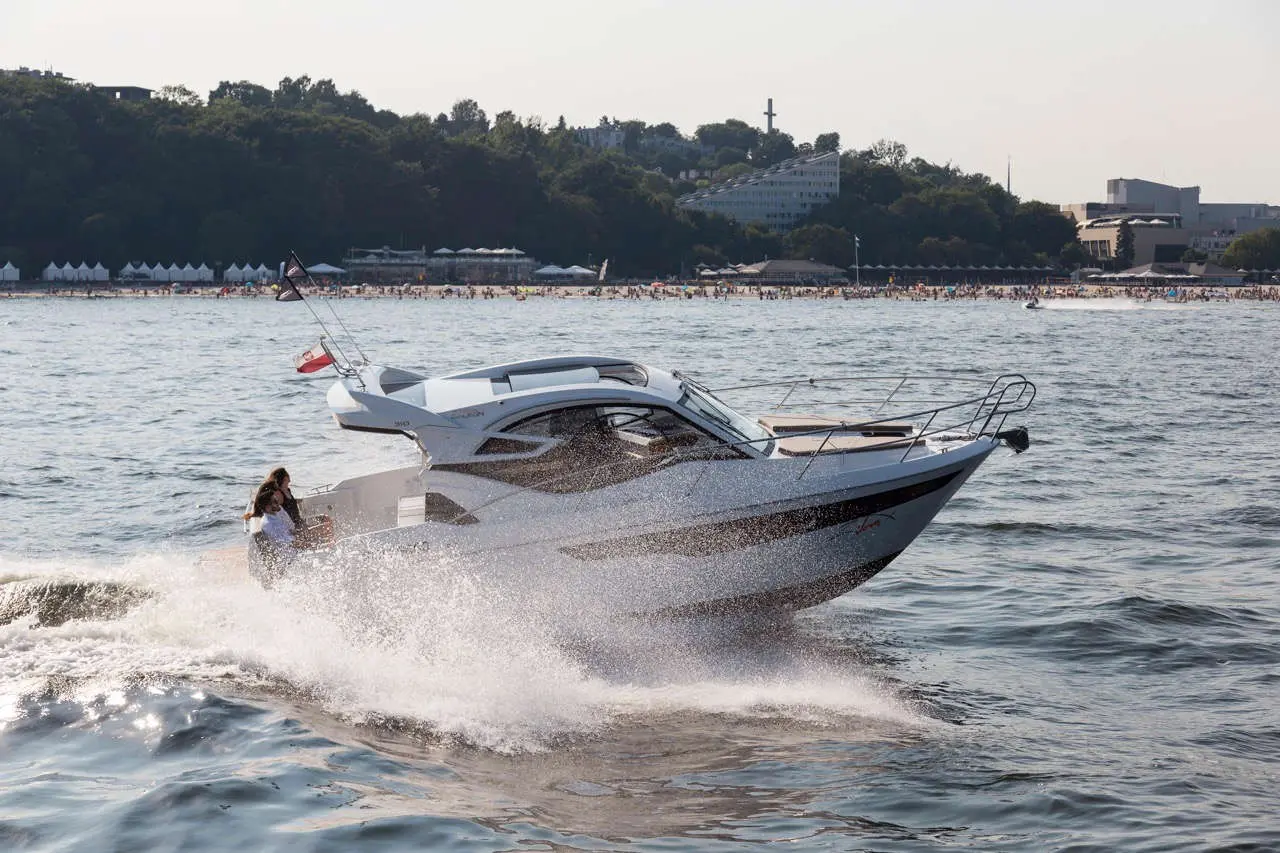 A sleek boat speeds through the water, creating a spray, with a beach and crowds in the background under a sunny sky.