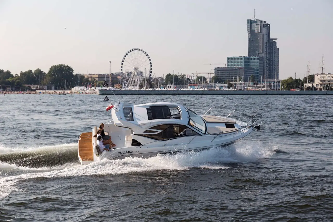 A white motorboat cruising on a river with two people aboard, a ferris wheel and city skyline in the background.