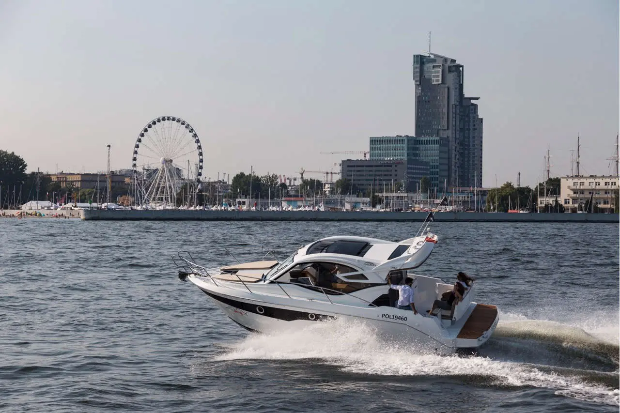 A white motorboat speeds across the water, with a Ferris wheel and modern buildings in the background.