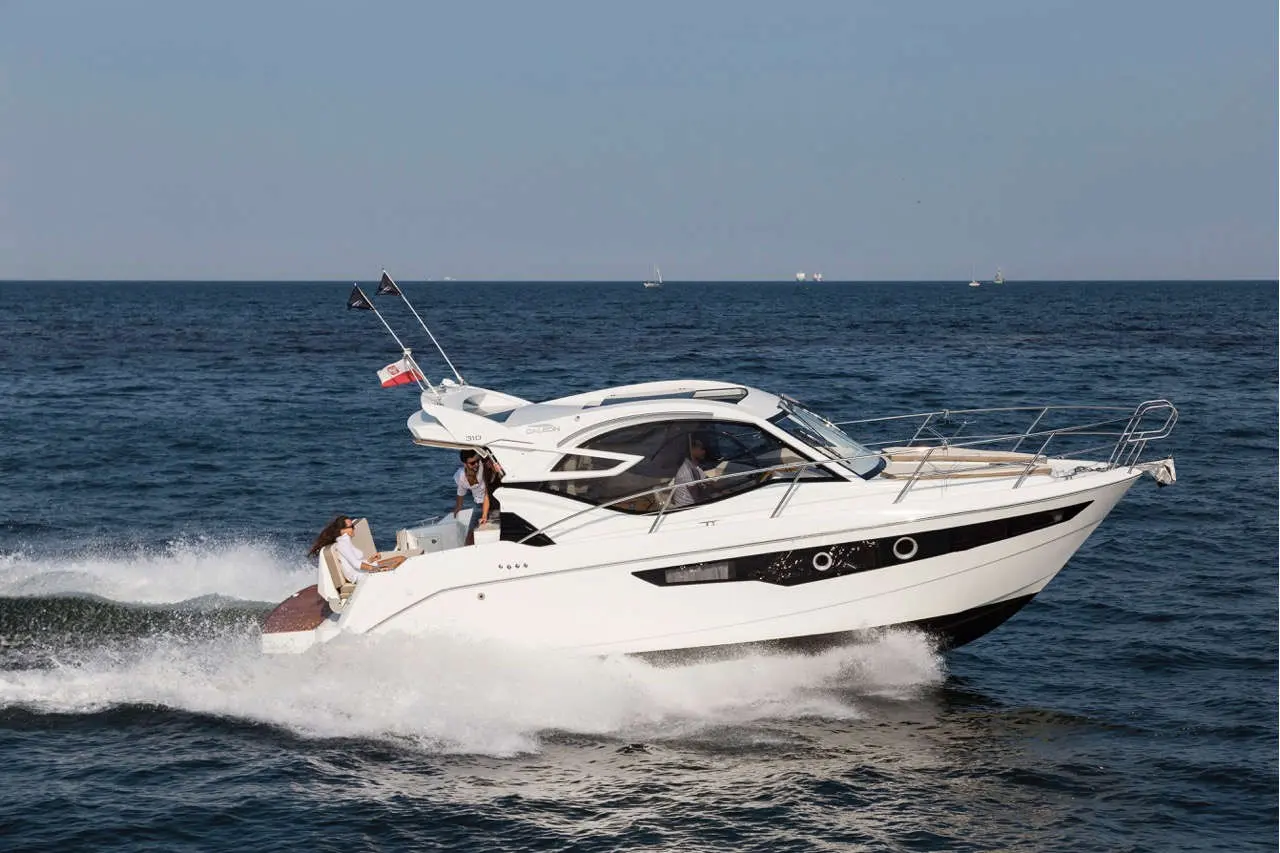 A white motorboat speeds across the ocean, two people on board enjoying the ride, with flags flying above.