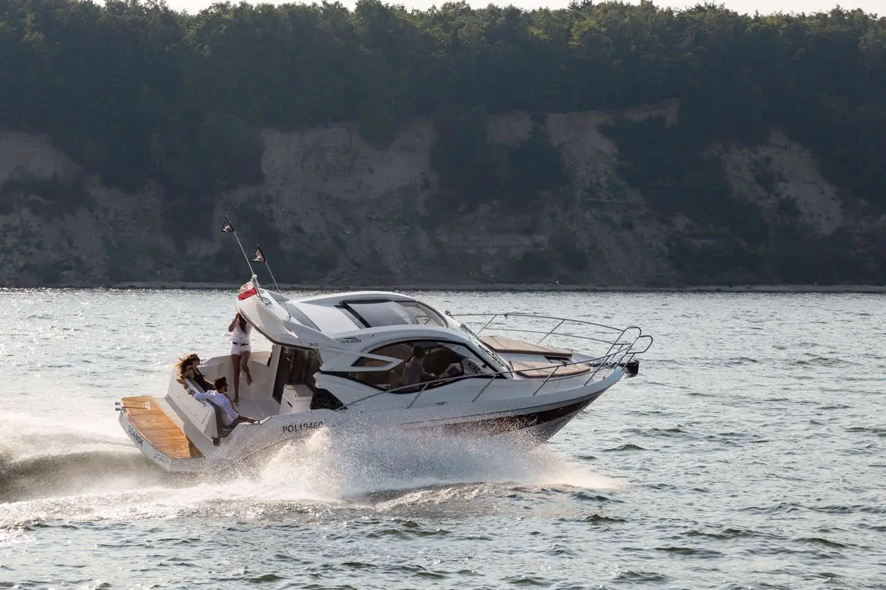A sleek motorboat speeds across the water, with two people enjoying the ride against a natural landscape.