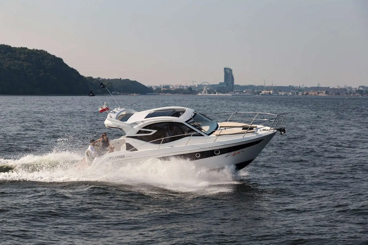 A white motorboat speeds across the water, creating spray, with a city skyline visible in the background.