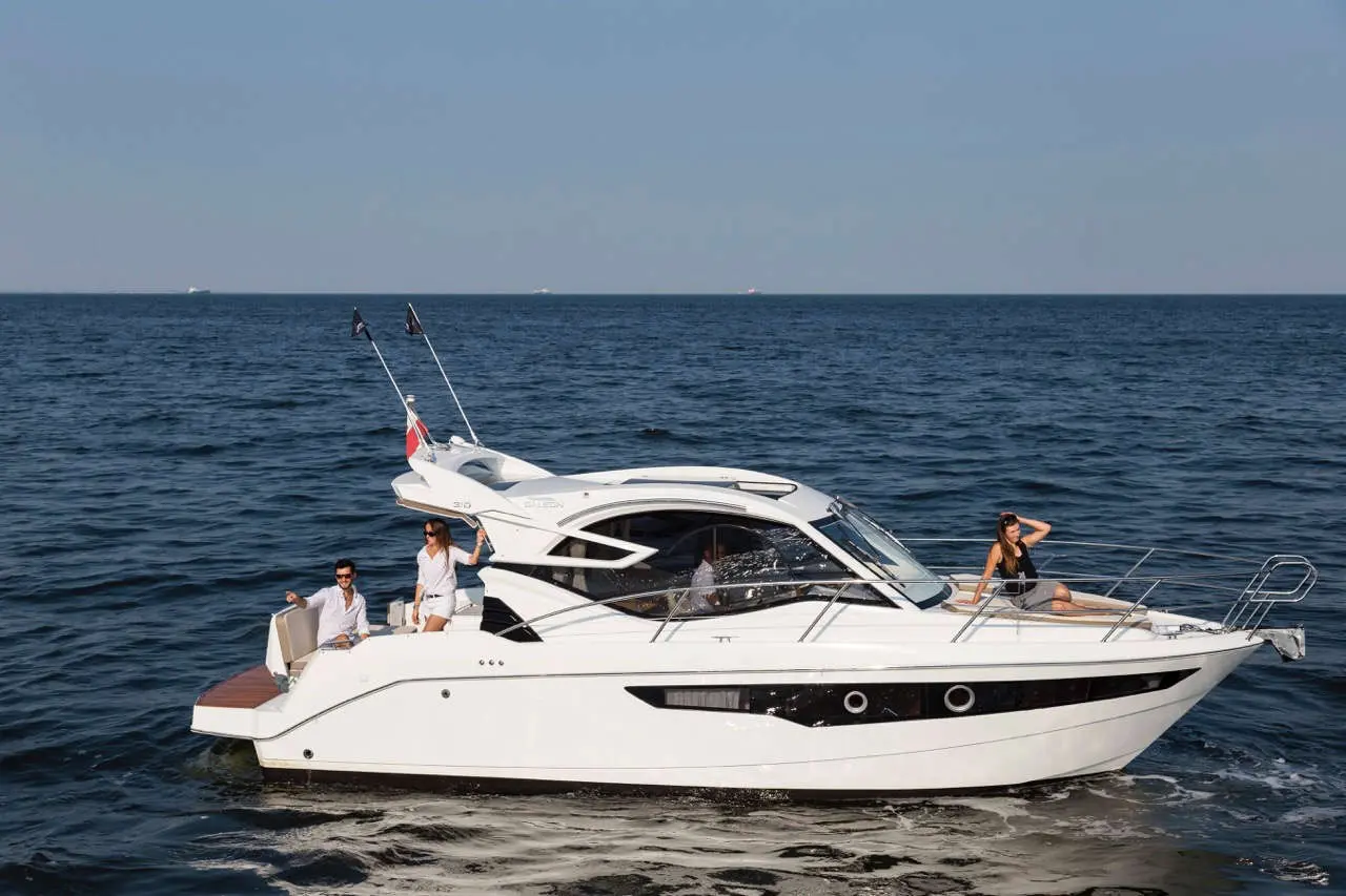 A modern white motorboat on a calm sea, with two people enjoying the sun and waving from the deck.