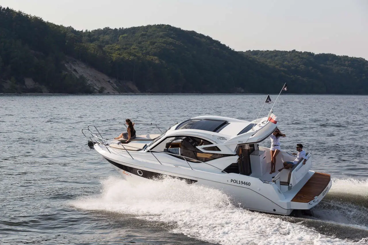 A luxury motorboat glides across the water, with two people enjoying the sun and stunning scenery of green hills.