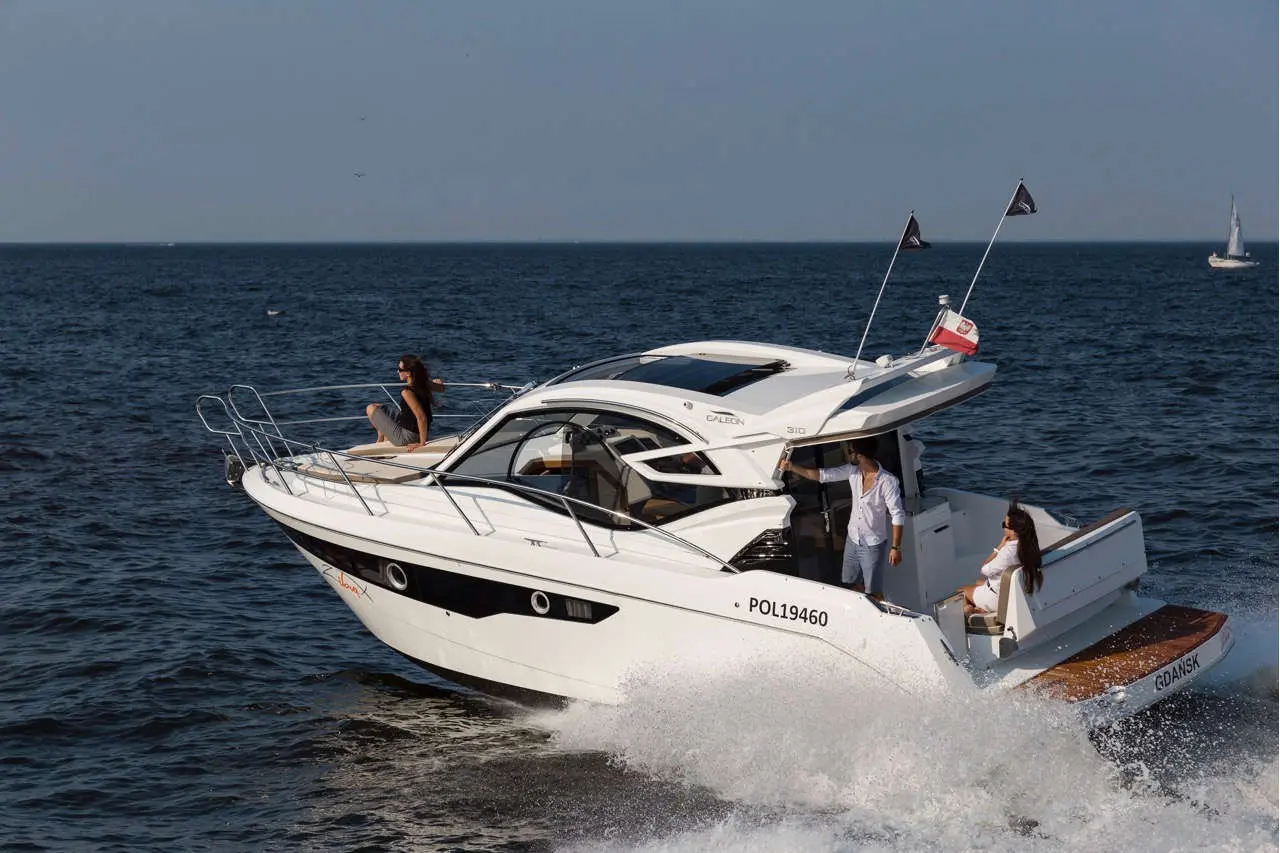A white motorboat cruising on water, carrying three people; one sitting at the bow and two at the stern, waves splashing.
