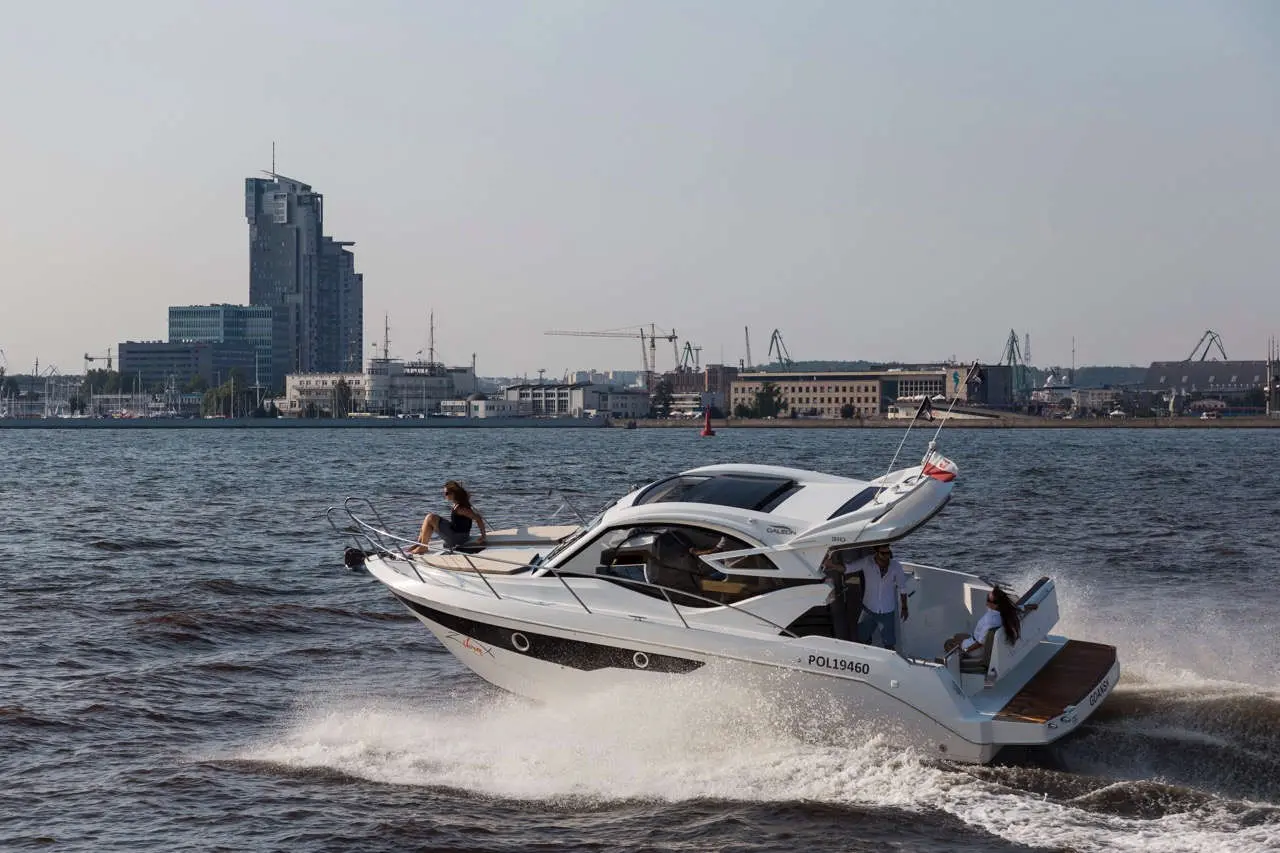 A white motorboat speeds across the water, with a city skyline in the background and two people onboard enjoying the ride.