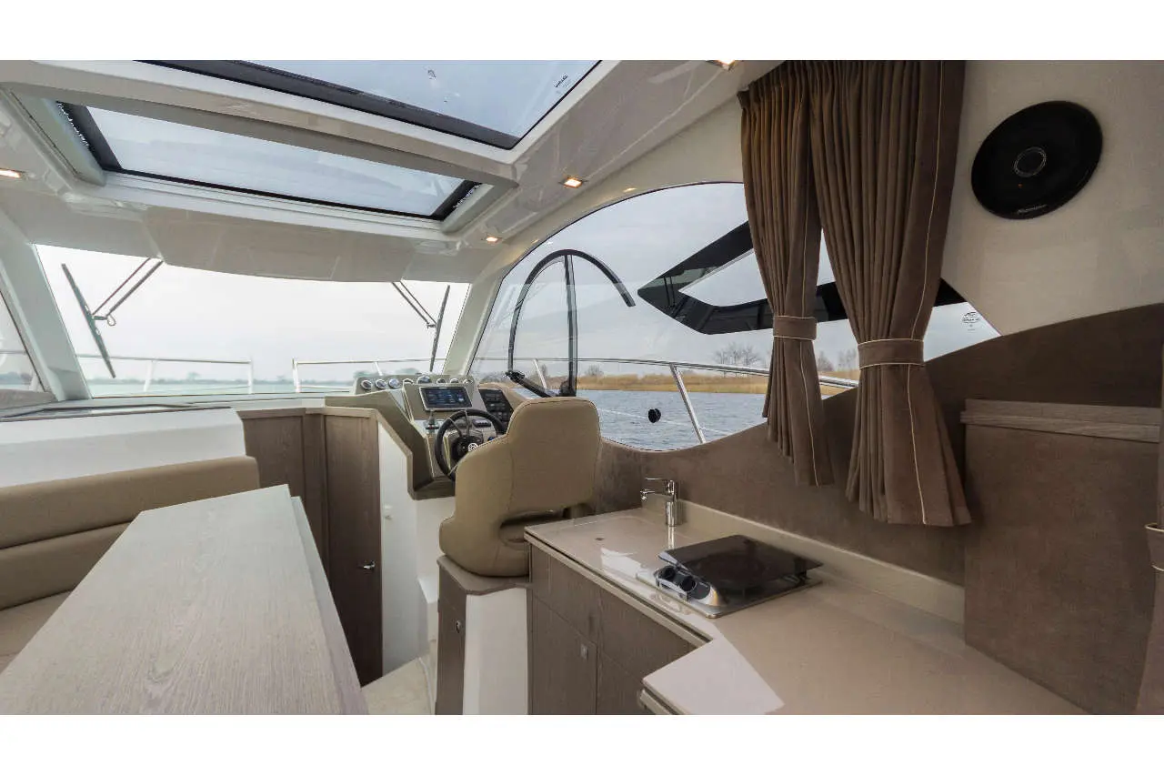 Interior of a modern boat featuring a steering wheel, control panel, and seating area with water view through large windows.
