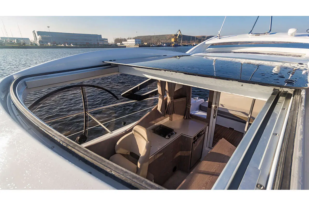 Interior view of a yacht showing a helm with a seat, a kitchenette, and large windows overlooking the water.