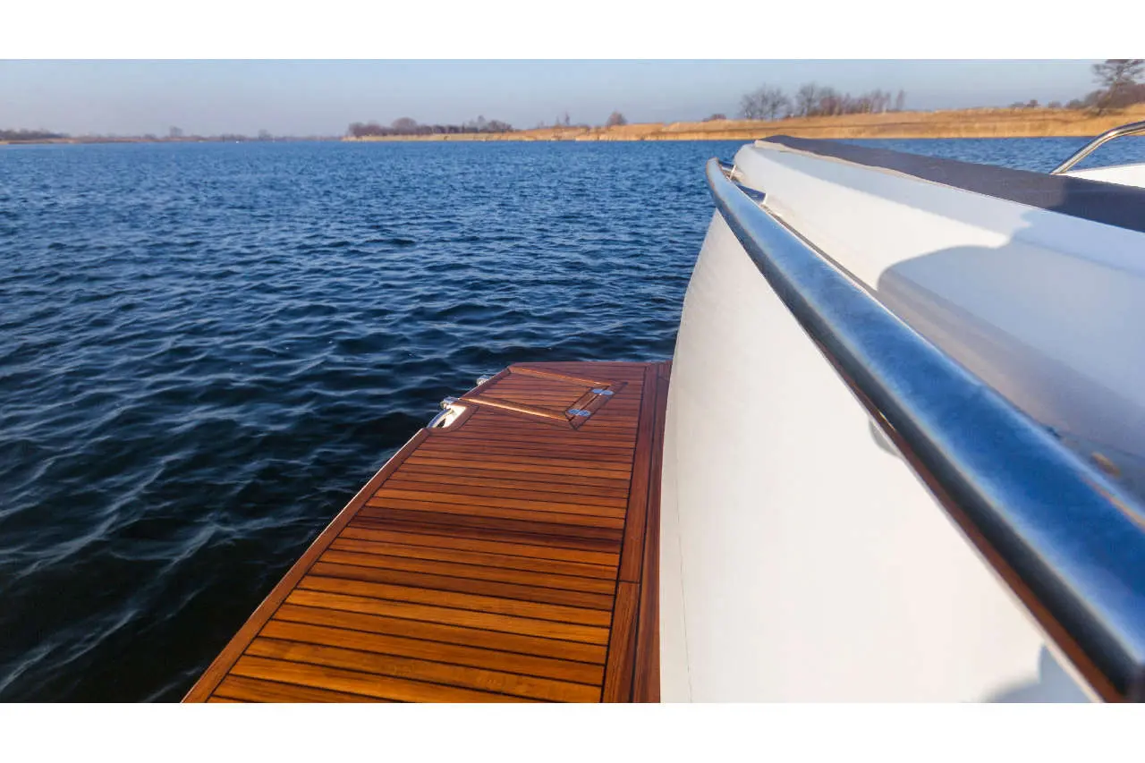 View from a boat showing polished wooden decking, stainless steel rail, and calm blue water under a clear sky.