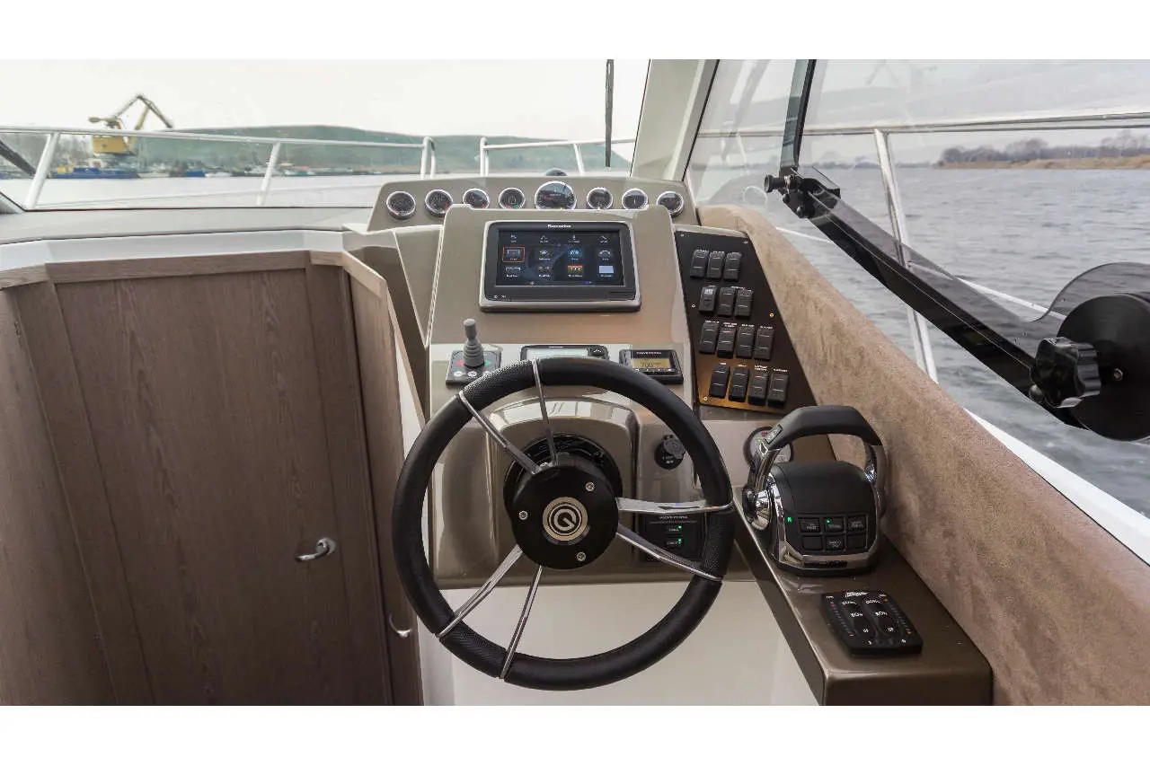 Boat dashboard featuring a steering wheel, control panel, display screen, and throttle controls, with water visible outside.