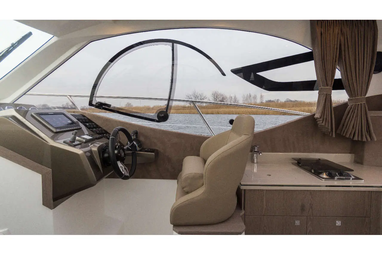 Luxury yacht interior with a steering wheel, control panel, and comfortable seating facing a scenic waterfront view.
