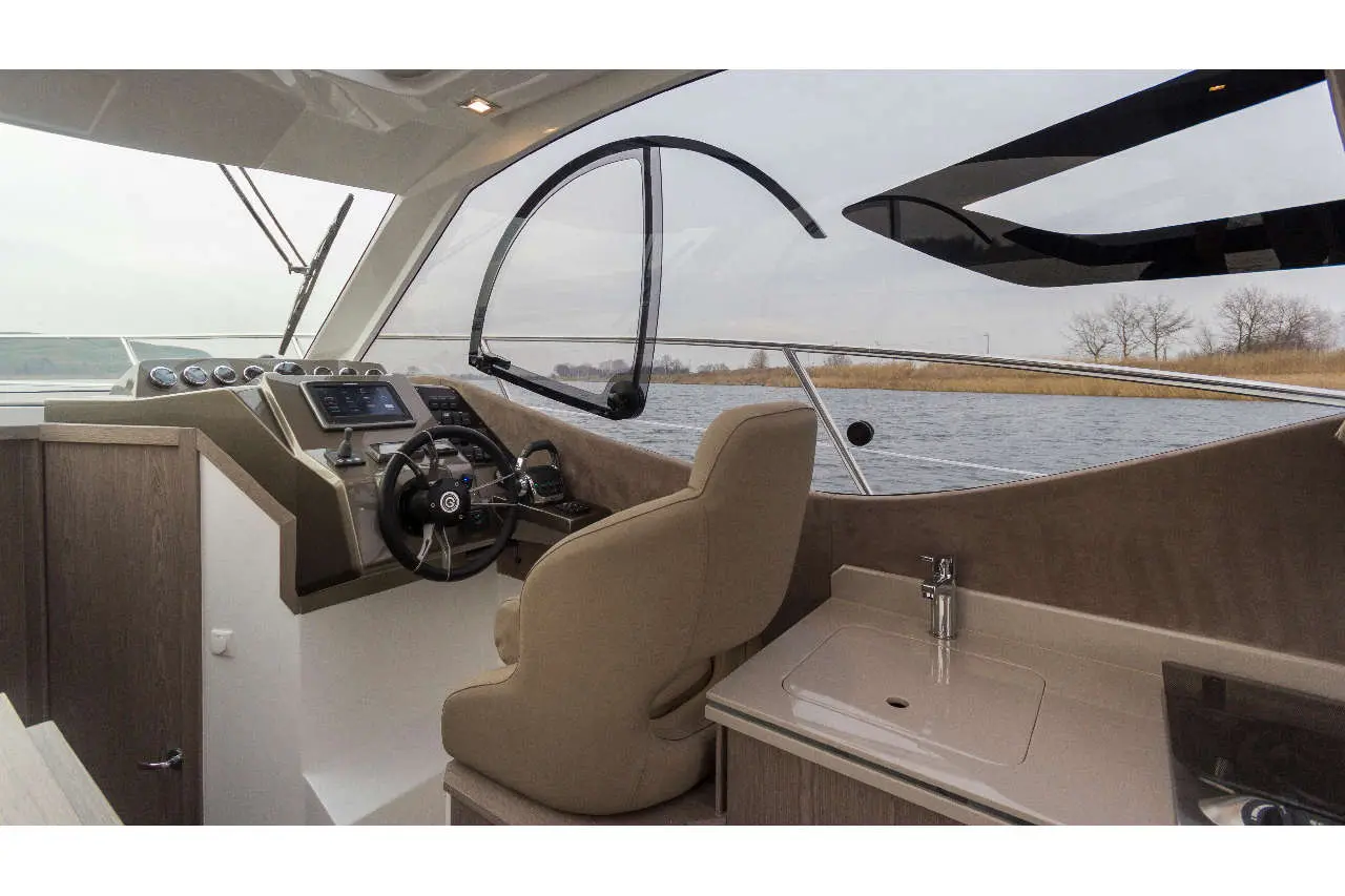 Interior view of a modern boat helm with steering wheel, control panel, and upholstered seat, overlooking water and landscape.