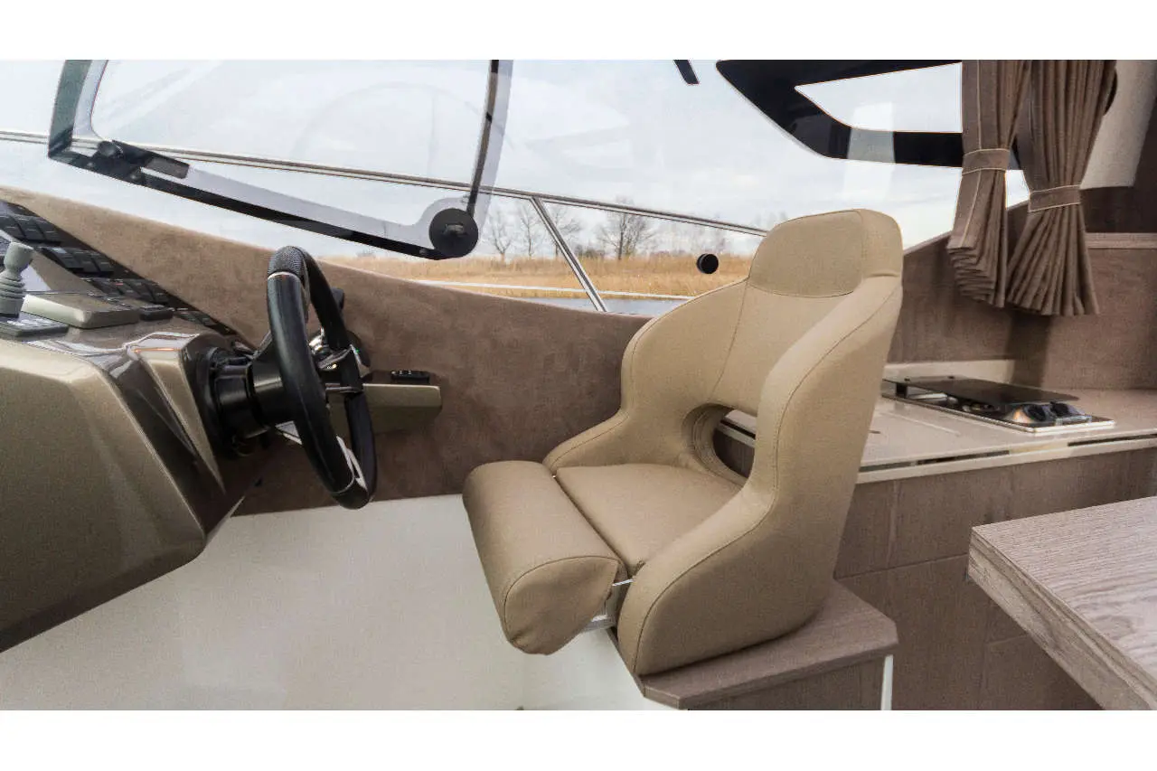 Interior of a yacht showing a beige helm chair and steering wheel, with a view of the water and cabinetry in the background.