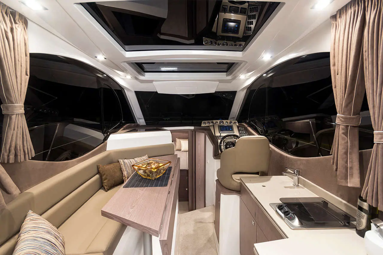 Modern yacht interior featuring a helm, seating area with cushions, and kitchenette, illuminated softly in low light.