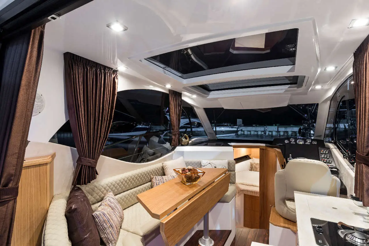 Modern yacht interior with a dining area, plush seating, wooden table, and a view of boats through large windows at night.
