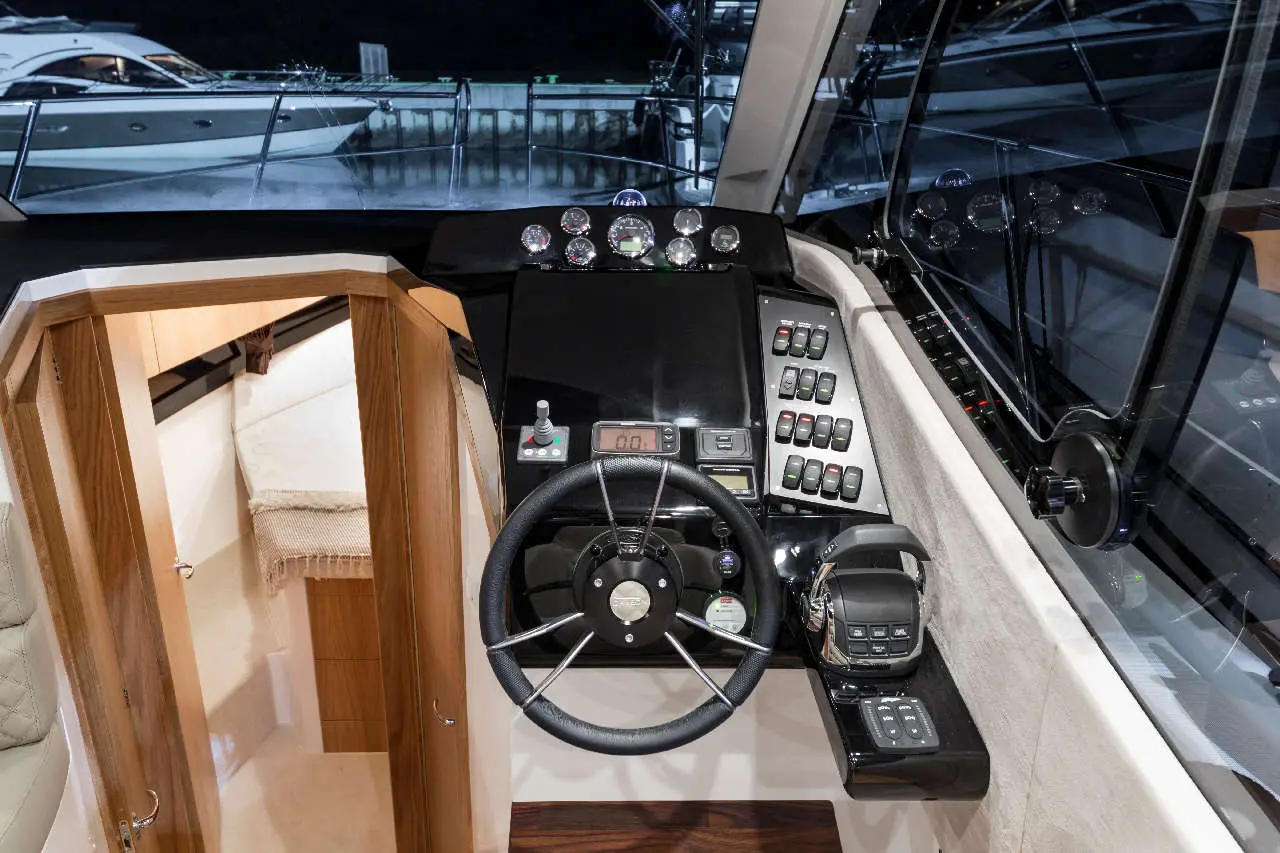 Boat cockpit interior featuring a steering wheel, control panel, gauges, and surrounding boats visible through the windows.