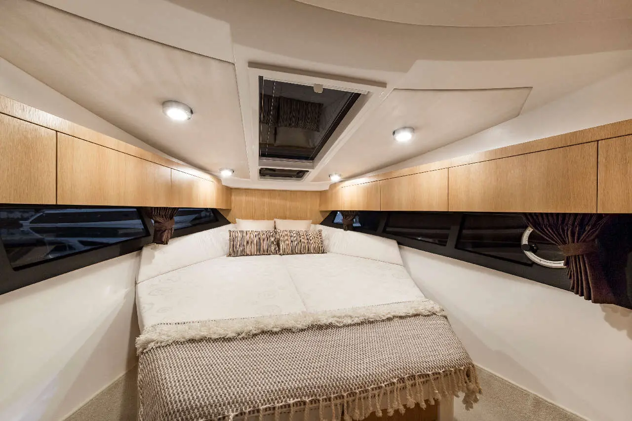 Cozy boat interior featuring a large padded bed, wooden storage cabinets, and windows with curtains, illuminated by ceiling lights.