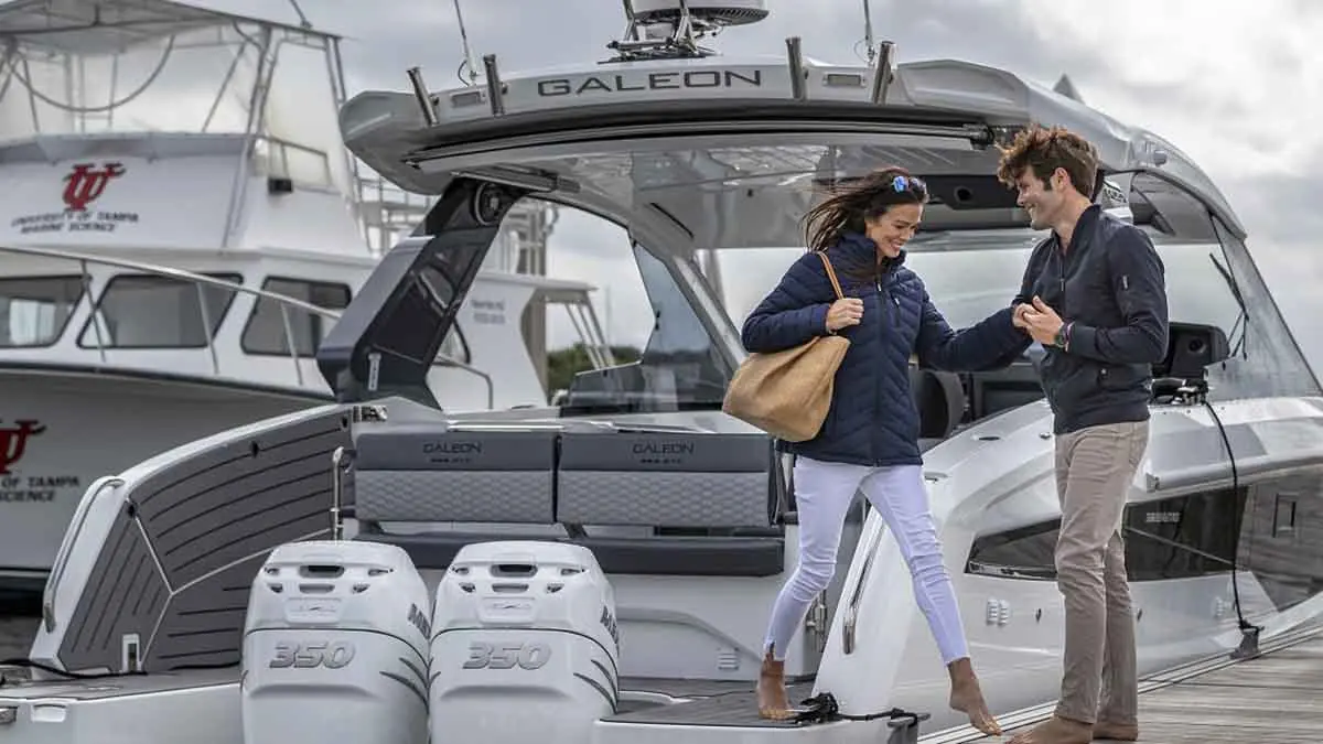 A woman and a man interact near a luxury Galeon boat, both dressed casually, with a marina in the background.